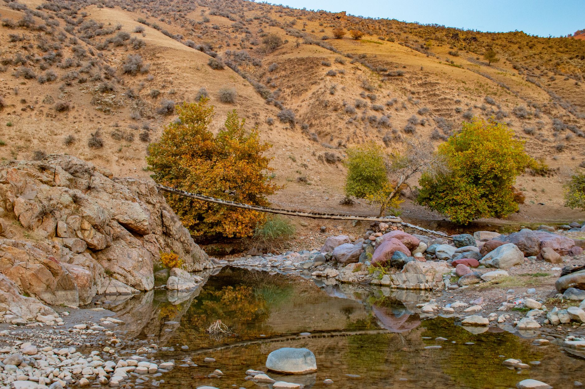 Scene depicting a desert valley with a stream reflecting autumn colors and a rustic rope bridge · Fergana Valley, Uzbekistan