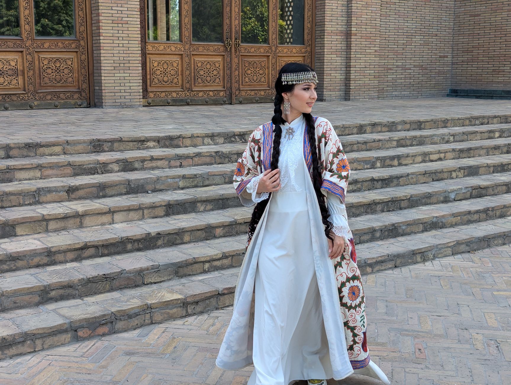 A woman in traditional Uzbek dress poses gracefully outside a historic building with detailed wooden doors, Uzbekistan