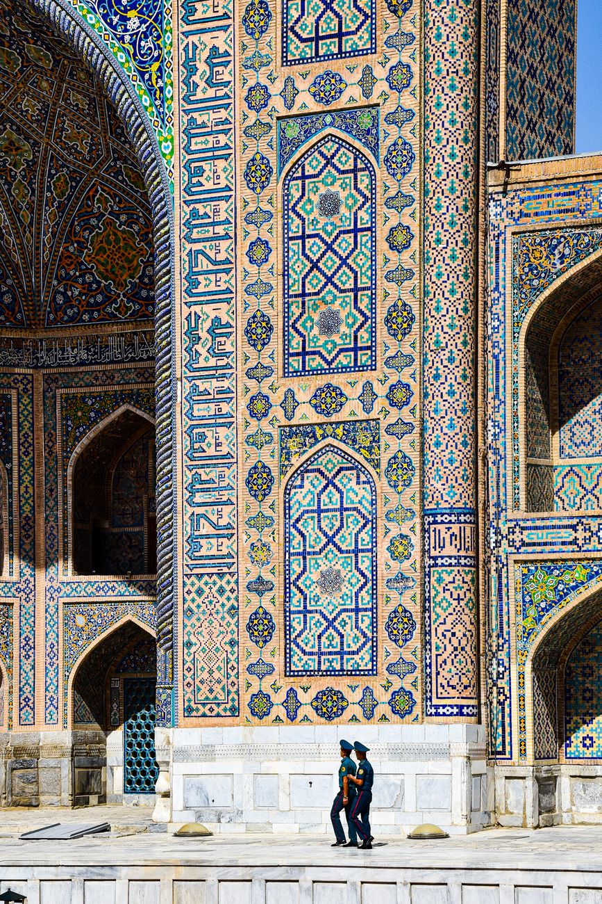Two men walk past the ornate facade of a historical building with intricate mosaics in Samarkand, Uzbekistan